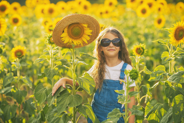 顽皮的女孩戴着黑色太阳镜，戴着草帽的向日葵，站在长满向日葵的田野里。阳光明媚的照片。图片下载