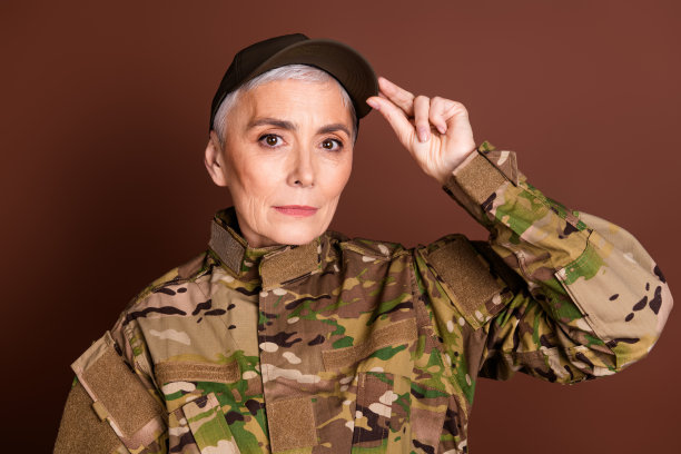 可爱的退休女士的照片肖像专业军官触摸帽子穿着迷彩军装孤立的棕色背景图片下载
