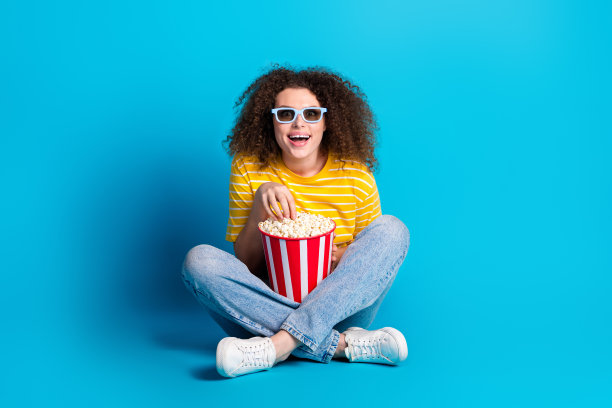 年轻女子的全身照片坐在地板上看电影吃爆米花穿着时尚的黄色条纹衣服孤立在蓝色背景图片下载