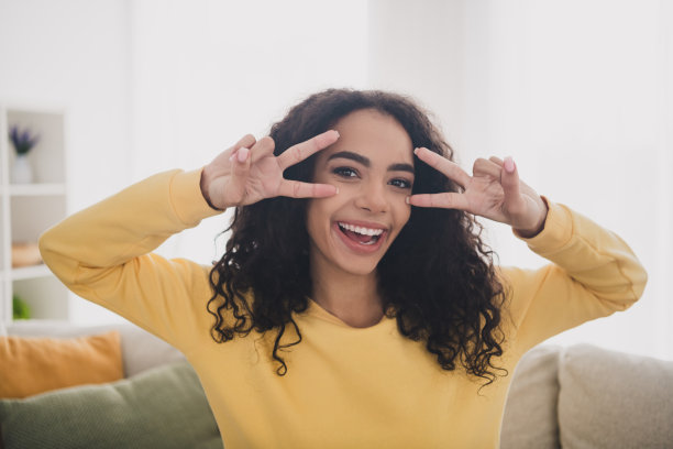 照片肖像，开朗的年轻女孩穿着黄色衬衫，乐观地摆出两个和平符号v标志在客厅里与日光图片下载