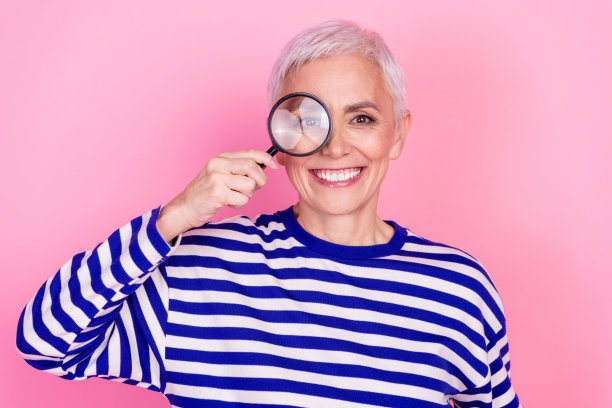 照片迷人可爱的高级妇女穿条纹时尚的衣服持有放大镜孤立在粉红色的背景图片下载
