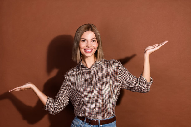 照片中漂亮可爱的女人穿着格子时尚的衣服，两只手臂抱着空旷的空间，孤立在棕色的背景上图片下载