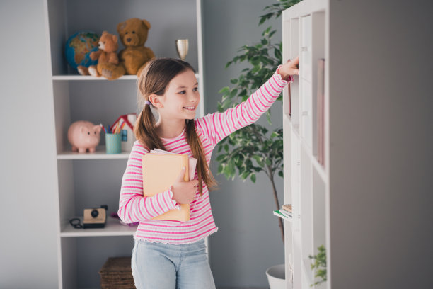 照片中可爱可爱的女孩女儿拿着图书馆书架上的书，从家里到室内远程上课教育图片下载