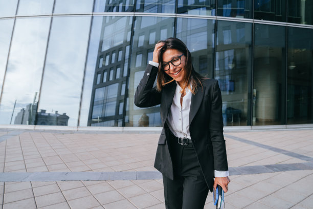 一位穿着黑色西装的职业女性微笑着整理着头发，走在一栋现代化的办公楼外面图片下载