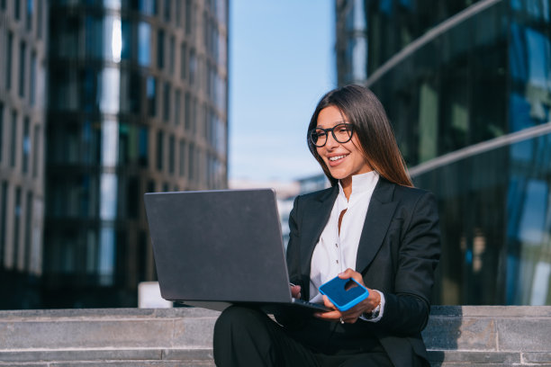 一位身着黑色西装、面带微笑的女商人在户外用笔记本电脑工作，手里拿着一部蓝色智能手机，背景是现代建筑图片下载