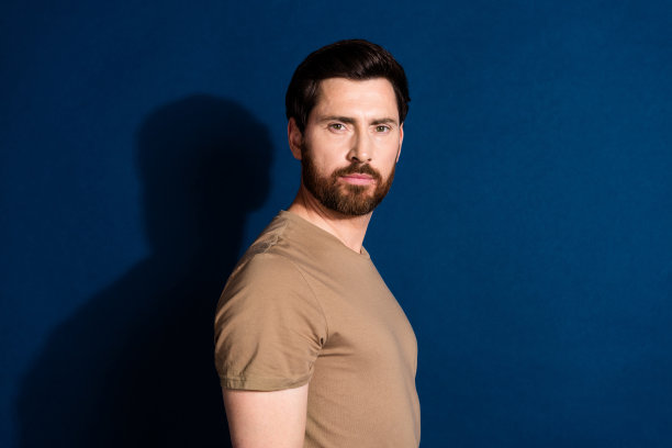 侧面资料照片好心情严肃的男人，留着胡子，头发，穿着米色t恤，看着你孤立的深蓝色背景图片下载