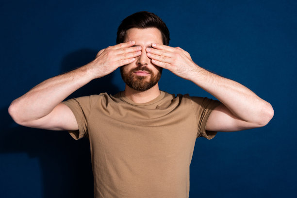 照片中的英俊严肃的男人，留着时髦的胡子，穿着米色t恤，手掌遮住眼睛，孤立在深蓝色的背景下图片下载