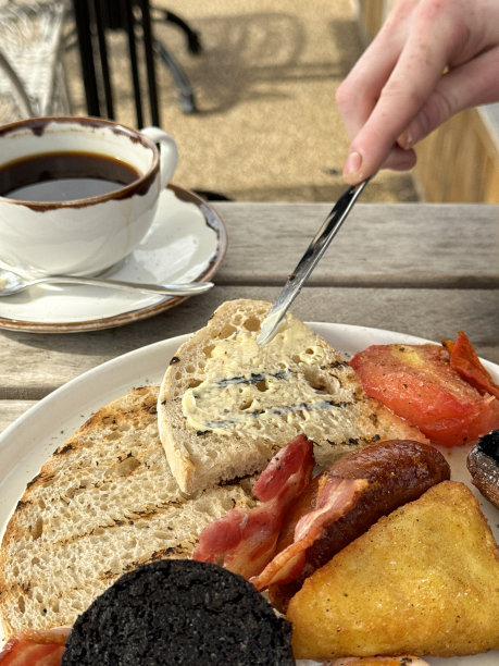 全英式早餐的特写图片，无法辨认的人用金属刀涂黄油白吐司，培根，香肠，烤西红柿，黑布丁，波多贝罗蘑菇，土豆泥，杯子和茶碟里的黑咖啡，餐馆的桌子图片下载