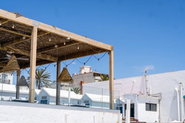 这张照片捕捉了一个宁静的夏日，展示了地中海建筑的魅力，白色的建筑，悬挂着灯的藤架，以及湛蓝天空下的稻草遮阳篷。图片下载