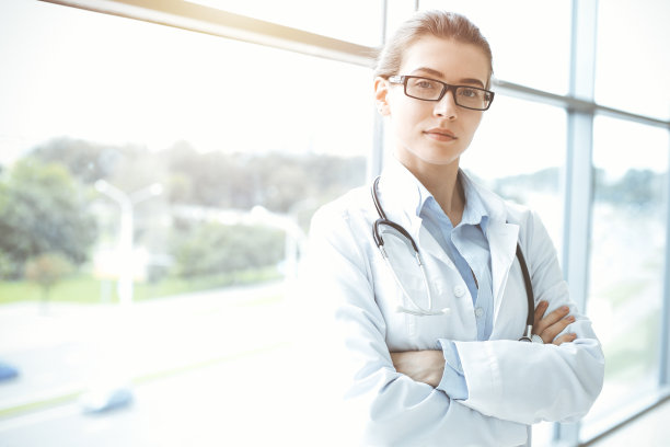 在阳光明媚的医院里，带着听诊器的美女医生微笑着，随时准备帮助病人。医学的概念图片下载