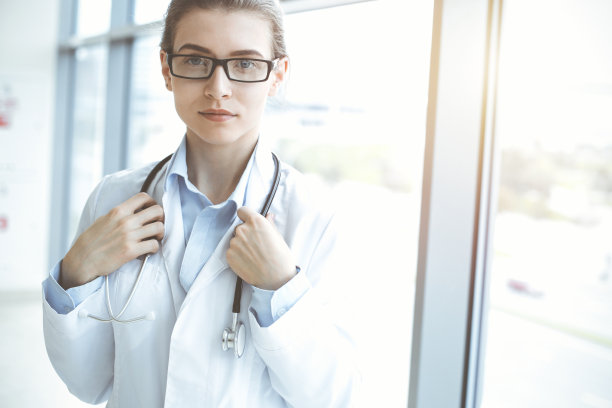 在阳光明媚的医院里，带着听诊器的美女医生微笑着，随时准备帮助病人。医学的概念图片下载