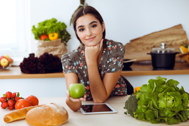 一位迷人的黑发年轻女子，手捧青苹果，面带微笑地在厨房里挑选美味佳肴的配方。平板电脑是最好的食谱图片下载