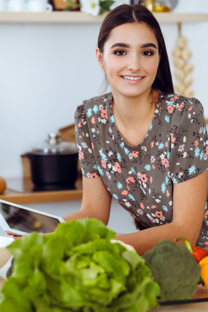 一位年轻迷人的黑发女子坐在厨房的餐桌旁，正在挑选一顿美味佳肴的食谱。平板电脑是最好的食谱图片下载