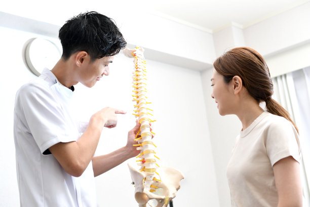 一位身穿白大褂的亚洲男性脊椎按摩师拿着脊椎模型向病人解释。图片下载