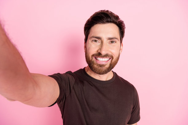 这张照片是一个长着牙齿、笑容灿烂的好男人，留着时髦的胡茬，穿着棕色t恤，在粉红色的背景下独自微笑着自拍图片下载