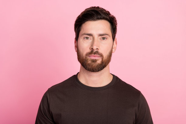 照片中，一个英俊、严肃、自信的男人，留着深色的胡茬，穿着棕色的t恤，在粉红色的背景下凝视着相机图片下载