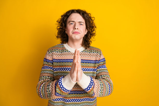 忧心忡忡的悲伤男子的照片，波浪发型，穿着装饰毛衣，祈祷，手掌放在一起，孤立在黄色背景上图片下载