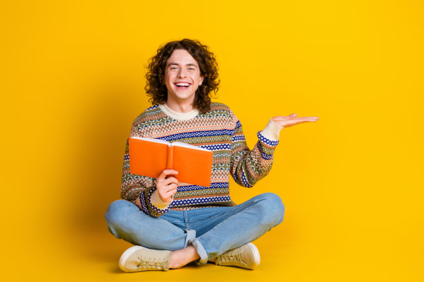 滑稽的家伙穿着装饰毛衣坐在地板上的全尺寸照片，拿着书手掌，在黄色背景上显示出孤立的空白空间图片下载