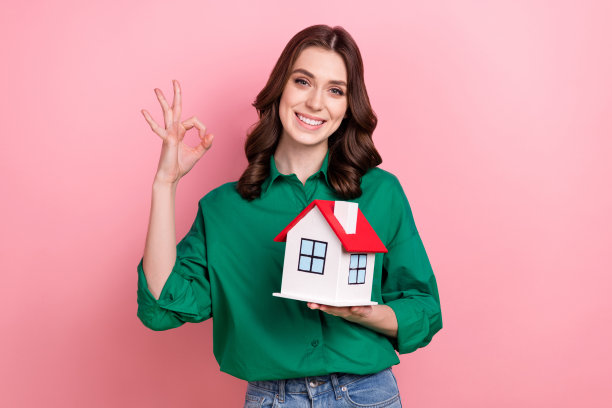 照片中，自信的漂亮女人穿着绿色衬衫，拿着小房子，显示出ok的标志，孤立的粉红色背景图片下载