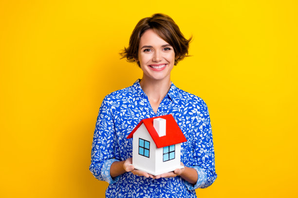 照片肖像漂亮的年轻女孩抱着小房子的房地产经纪人穿着时尚的印花服装孤立在黄色的背景图片下载