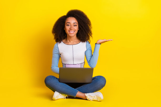 漂亮的年轻女孩的全尺寸照片，坐在地板上上网本，拿着空白的空间，穿着时髦的蓝色衣服，孤立在黄色的背景上图片下载