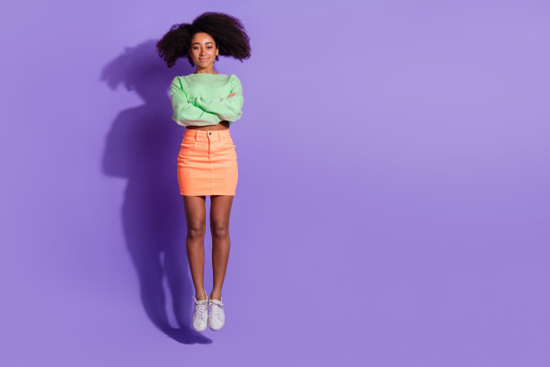 年轻有趣的跳跃折叠双手女孩的全身长度的照片，波浪的头发在绿色的顶部和橙色的迷你裙孤立的紫色背景图片下载