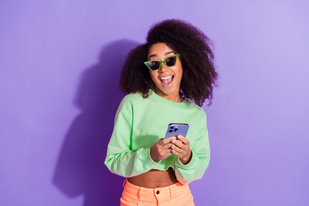 照片中的美国年轻开朗有趣的女人戴着时髦的太阳镜，在紫色的背景下用智能手机跟踪社交网络图片下载