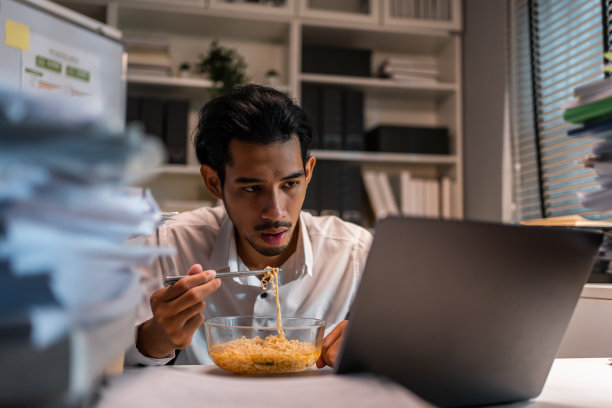 亚洲年轻商人晚上在办公室工作时吃面条。有吸引力的职业男性员工坐在桌子上，感到饥饿和饥饿，然后在公司工作场所吃晚餐。图片下载