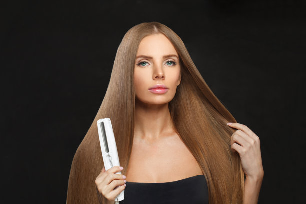 名人黑发女子拿着直发棒，在白色背景上拉直她光滑闪亮的长发。护发、发型、美发及发型设计理念图片下载