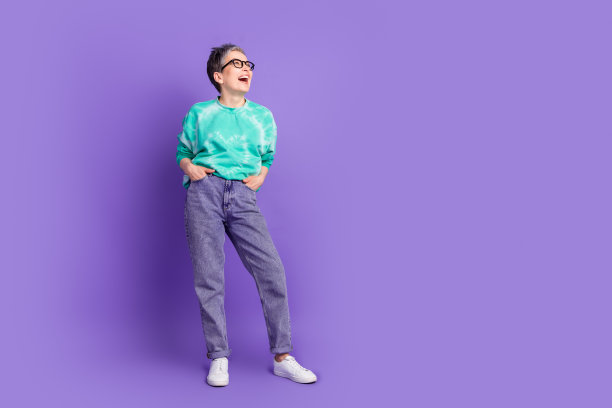 照片中的资深女性笑着摆姿势，双手口袋看起来空空，穿着绿色的衣服，孤立在紫色的背景上图片下载