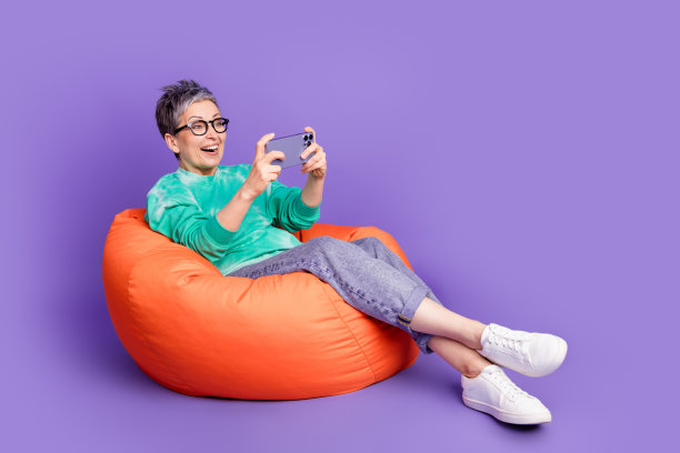 迷人的老年妇女全身照片坐在豆袋玩手机游戏穿着时尚的绿色衣服孤立在紫色的背景图片下载