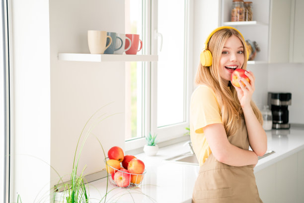 漂亮的十几岁的女孩的照片耳机听音乐吃苹果穿围裙烹饪美味的饭菜食谱厨房室内图片下载