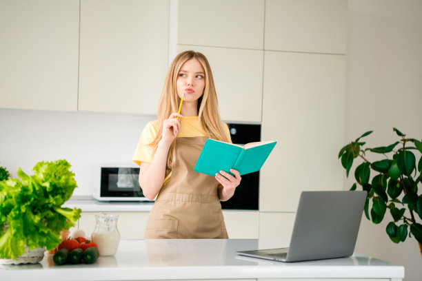 漂亮的十几岁的女孩的照片看起来头脑混乱的食谱写笔记本电脑笔记本电脑穿围裙烹饪美味的饭菜食谱厨房室内图片下载