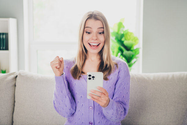 迷人的年轻女子的照片，坐在沙发上，拿着获胜的装置，穿着紫色的衣服舒适的日光家庭室内客厅图片下载