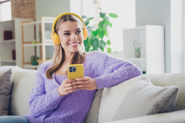 年轻女孩的照片，坐在沙发上，耳机，听音乐，拿着设备，看空空间，穿紫色的衣服，现代舒适的室内，平坦的室内图片下载