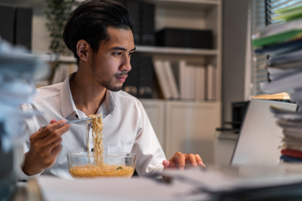 亚洲年轻商人晚上在办公室工作时吃面条。有吸引力的职业男性员工坐在桌子上，感到饥饿和饥饿，然后在公司工作场所吃晚餐。图片下载
