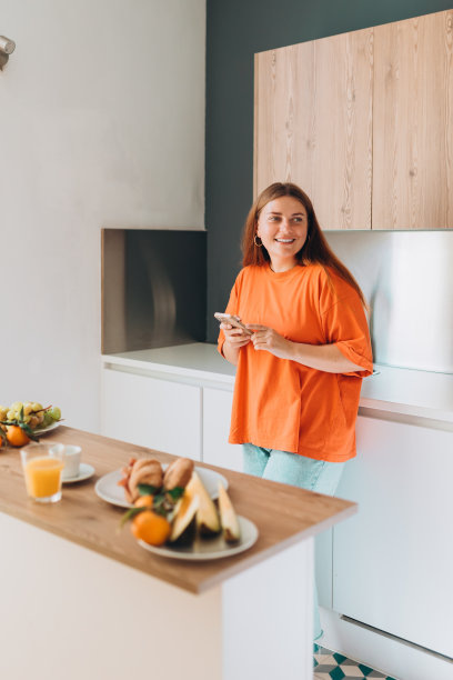 一个30多岁的女人站在厨房里，手里拿着手机。家庭生活和休闲时光。高质量的照片。图片下载