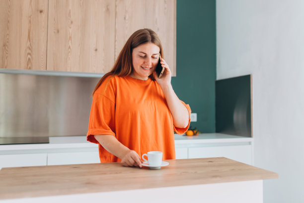 在慵懒的早晨，在舒适的厨房里，悠闲随意的年轻美丽的快乐微笑的女人喝着咖啡。30多岁的女人在家里打电话。高质量照片图片下载
