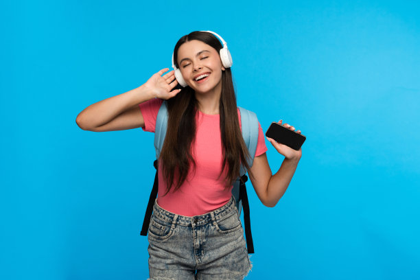 年轻的女孩小学生用智能手机听着蓝色背景的耳机音乐。图片下载