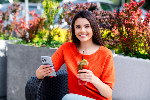 漂亮的青少年照片好心情的女孩坐在早餐喝鸡尾酒拿着装置穿着红色的衣服走在城市公园外面图片下载