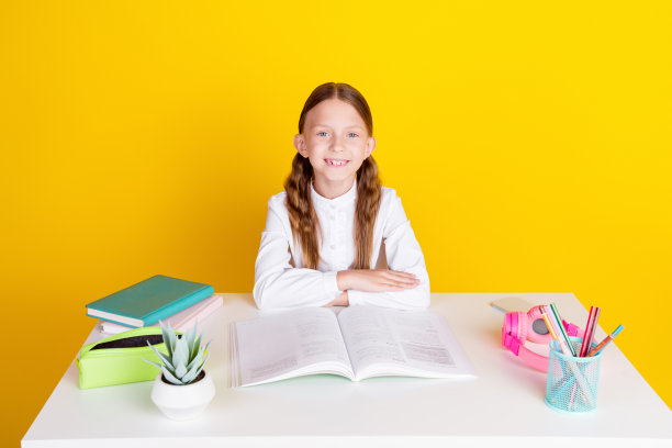 照片肖像可爱的小女生勤奋坐在桌子上穿着时尚的制服孤立在黄色的背景图片下载