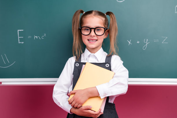 照片中可爱的小女生勤奋地拿着书本，穿着时髦的校服教室黑板背景图片下载