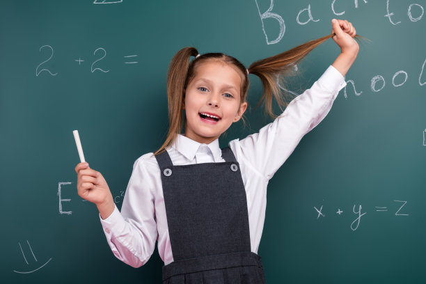 照片中迷人的小女生手持粉笔拉发尾，穿着时髦的校服教室黑板背景图片下载