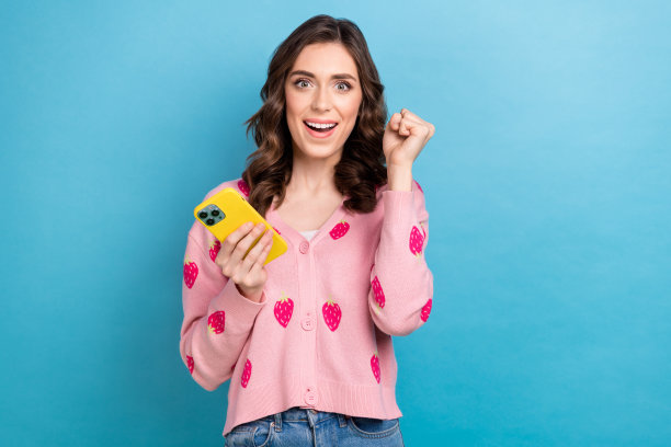 照片肖像的迷人的女人拿着电话，举起拳头，穿着时髦的粉红色草莓印花服装孤立的蓝色背景图片下载