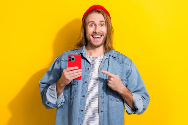 照片正面兴奋的男子穿着牛仔夹克指向苹果三星现代小工具孤立的黄色背景图片下载