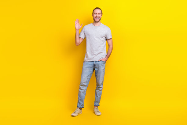 全尺寸照片的好心情友好的男人穿灰色t恤牛仔裤手在口袋里挥舞手掌说你好孤立的黄色背景图片下载
