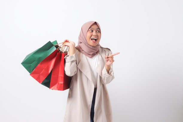 穿着休闲套装，戴着头巾的亚洲女性兴奋地拿着纸质购物袋，用手指着。购物狂和折扣购物概念。白色背景上的孤立图像图片下载