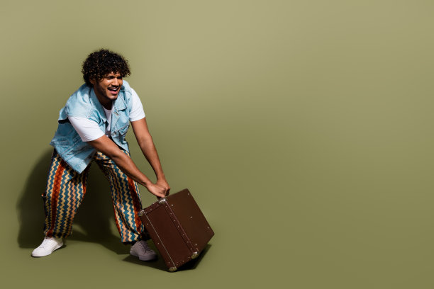 全尺寸照片，英俊的年轻人拉着沉重的复古行李箱，穿着时髦的牛仔裤，以卡其色为背景图片下载