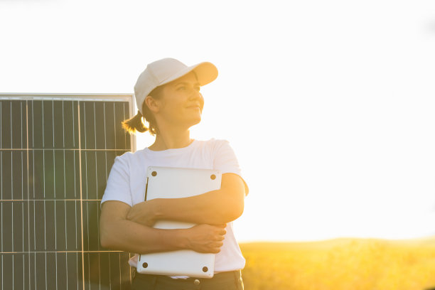 戴着白色帽子、穿着t恤、拿着笔记本电脑的农妇站在太阳能电池板旁边图片下载