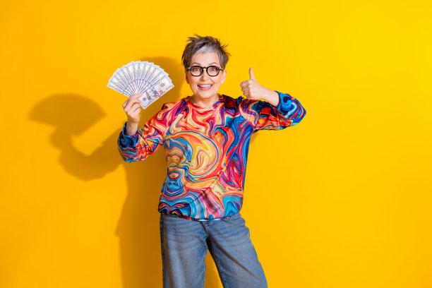 照片中，漂亮的老妇手拿钱，大拇指朝上，穿着黄色背景的套头衫图片下载